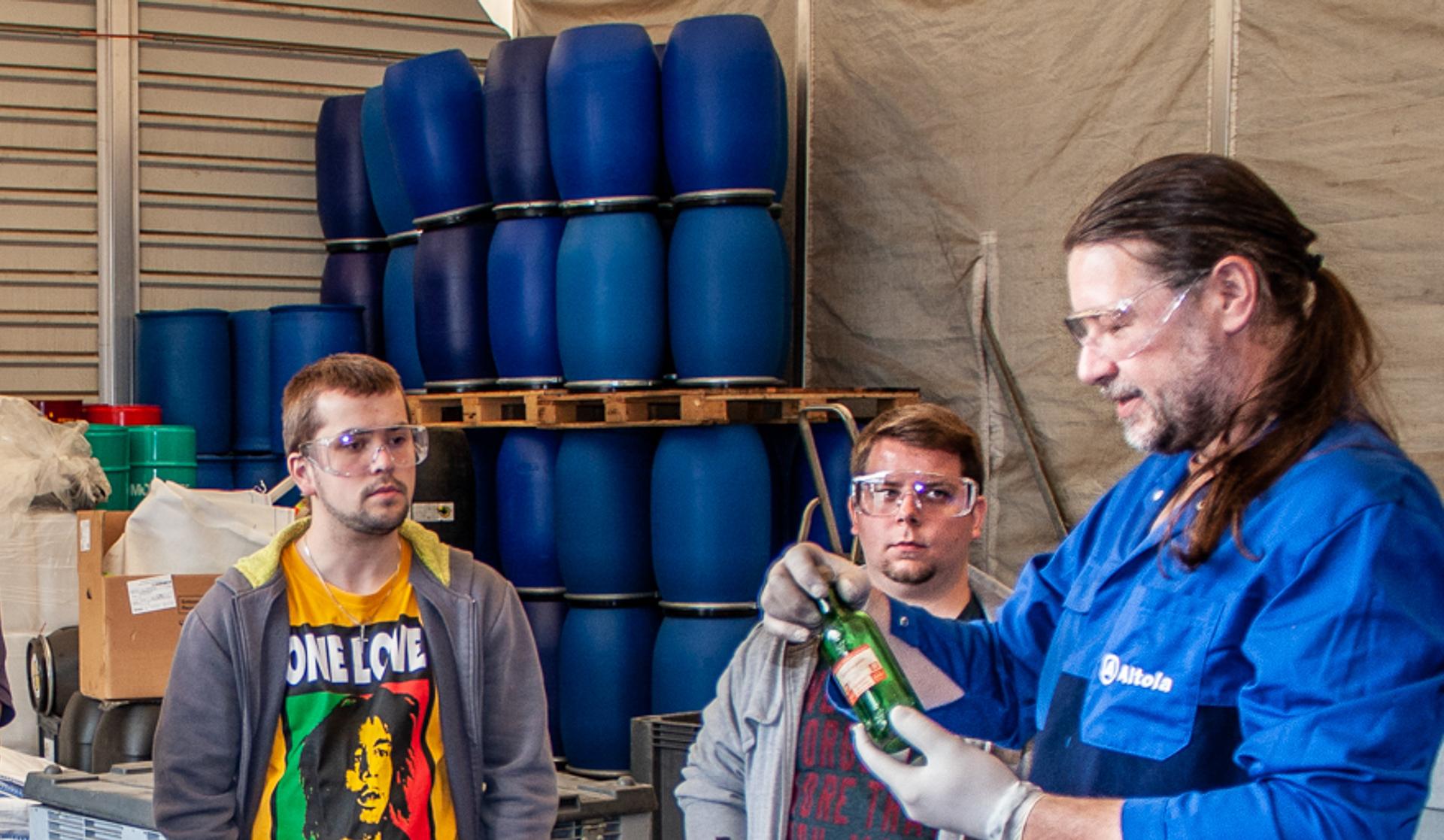 Der Kursleiter von Altola in der Sicherheitsausrüstung erklärt anhand einer Glasflasche, die er in den Händen hält die Gefahr des Inhaltsstoffes.